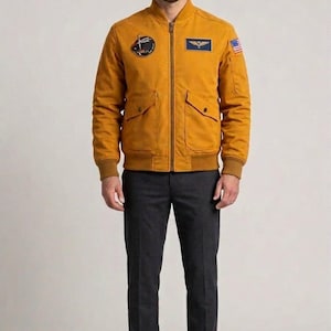 May include: Mustard yellow bomber jacket with a zip closure, two front pockets, and ribbed cuffs. Features patches on the chest and arm, along with dark grey trousers. The jacket has an American flag patch.