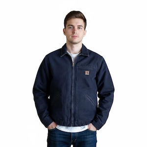 Op de afbeelding: Een donkerblauwe spijkerjas met een ritssluiting over de hele lengte en een kleine bruine logopatch op de borst. De jas heeft een kraag en twee voorzakken. De persoon draagt een wit T-shirt en een blauwe jeans. Klassieke workwear stijl.