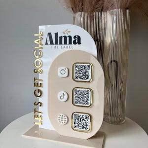 Könnte beinhalten: Ein beigefarbener und weißer Social-Media-Display-Ständer mit goldenen Akzenten. Der Ständer zeigt den Text "Alma THE LABEL" und "LET'S GET SOCIAL" in Gold. Er enthält QR-Codes für Instagram, TikTok und eine Website.