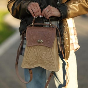 May include: A brown and tan leather backpack with a flap closure. The top flap is embossed with a bird and wheat design. The lower portion features a wheat and bird design. The bag has adjustable straps and a top handle.