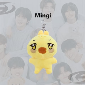 May include: A yellow plush keychain in the shape of a cartoon chick with a sad expression. The keychain has a silver clasp and the word "Mingi" is printed above the plush toy. The background features a group of people.