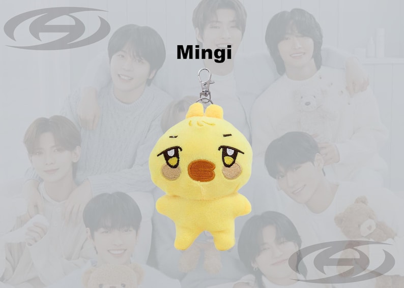 May include: A yellow plush keychain in the shape of a cartoon chick with a sad expression. The keychain has a silver clasp and the word "Mingi" is printed above the plush toy. The background features a group of people.