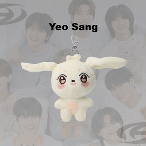 May include: A cream-colored plush bunny keychain with long ears, a pink nose, and embroidered eyes and mouth. The keychain is attached to a silver clasp. The background features a group of people.