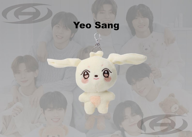 May include: A cream-colored plush bunny keychain with long ears, a pink nose, and embroidered eyes and mouth. The keychain is attached to a silver clasp. The background features a group of people.