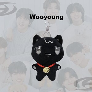 May include: Black plush cat keychain with a red collar and gold bell. The cat has white accents and large eyes. The word "Wooyoung" is above the cat. The background features a group of people.