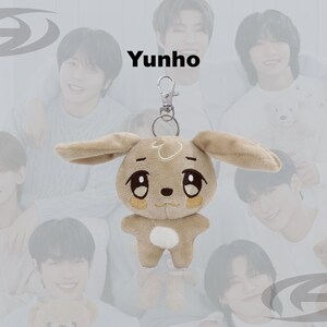 May include: A plush keychain featuring a tan bunny with large eyes and long ears. The keychain has a silver clasp and the word "Yunho" is printed above the plush toy. The background features a group of people.