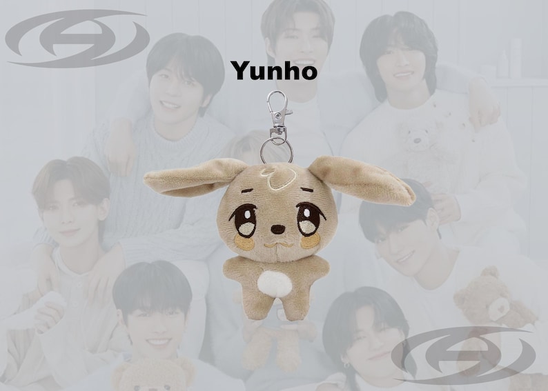 May include: A plush keychain featuring a tan bunny with large eyes and long ears. The keychain has a silver clasp and the word "Yunho" is printed above the plush toy. The background features a group of people.