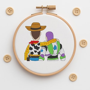 Peut inclure: Broderie sur tambour représentant deux personnages de dessins animés, l'un en costume de cow-boy et l'autre en combinaison spatiale, s'embrassant. Le motif est sur fond blanc, avec des boutons en bois éparpillés.