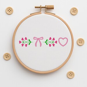 Pode incluir: Um bastidor de bordado de madeira com um tecido branco com um laço rosa, corações rosa e folhas verdes. O design é rodeado por botões de madeira.