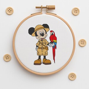 May include: Embroidered hoop art showing Mickey Mouse in safari clothing, with a red, yellow, and blue parrot. The design is set against a white background within a wooden embroidery hoop. Several wooden buttons are scattered around the hoop.