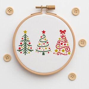 Pode incluir: Um bastidor de bordado de madeira com um tecido branco com três desenhos de árvores de Natal. As árvores são verdes, vermelhas e douradas, com detalhes de estrelas e ornamentos. Botões de madeira circundam o bastidor.