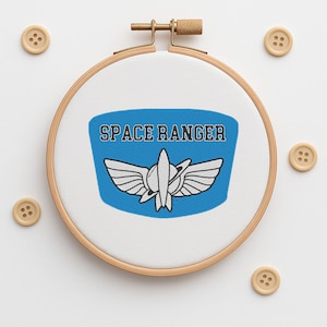 Pode incluir: Uma peça de arte bordada em bastidor com um emblema azul Space Ranger, texto branco e um desenho de foguete. O bordado está num bastidor de madeira, com quatro botões de madeira à volta. O bastidor tem cerca de 15 cm de diâmetro.
