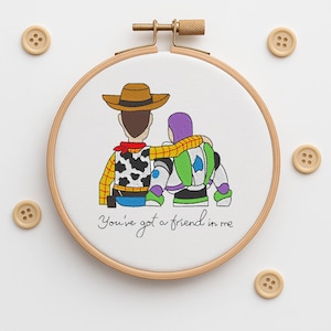 Pode incluir: Arte de bordado em bastidor com Woody e Buzz Lightyear abraçados, com o texto "You've got a friend in me". A obra de arte está em um bastidor de madeira, com quatro botões de madeira ao redor.
