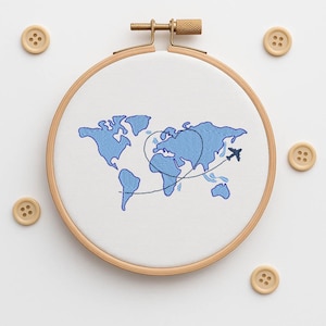 May include: An embroidery hoop with a world map design in blue thread. A small airplane is stitched near Australia, with a dotted line showing a flight path. The wooden hoop is surrounded by four wooden buttons.
