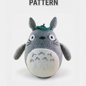 May include: A crocheted Totoro amigurumi pattern is displayed. The plush toy is gray with a white belly and a green leaf on its head. The text "TOTORO AMIGURUMI PATTERN" is at the top of the image.