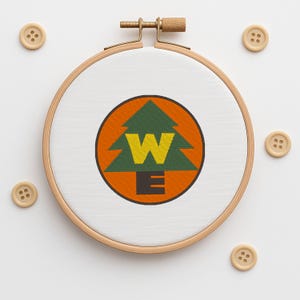 Pode incluir: Arte de bordado em bastidor com um desenho de árvore verde, a letra "W" amarela e a letra "E" preta dentro de um círculo laranja. O desenho está em tecido branco, emoldurado por um bastidor de madeira. Vários botões de madeira estão espalhados ao redor do bastidor.