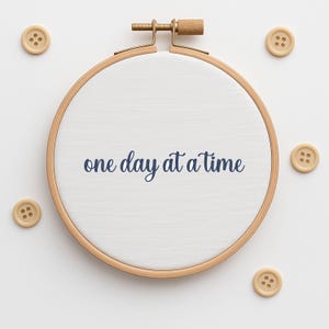 Pode incluir: Um bastidor de bordado de madeira com a frase "one day at a time" em azul marinho. Seis pequenos botões de madeira de cor clara circundam o bastidor, criando um design simples e elegante.