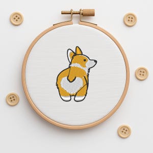 Peut inclure: Un cercle à broder avec un motif brodé de chien corgi. Le corgi est jaune et blanc, avec un contour noir. Plusieurs boutons en bois sont dispersés autour du cercle.