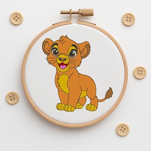 May include: An embroidery hoop featuring a cartoon lion cub. The lion cub is depicted in orange and yellow with a pink tongue. Wooden buttons are scattered around the hoop.