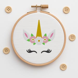 Pode incluir: Um bastidor de bordado de tecido branco com um desenho de rosto de unicórnio. O unicórnio tem um chifre dourado, orelhas rosa e brancas e uma coroa floral com flores rosa, verdes e brancas. Botões de madeira circundam o bastidor.