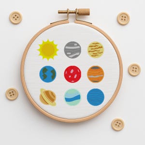 May include: A wooden embroidery hoop with a white fabric featuring embroidered planets and a yellow sun. The planets are in various colors, including blue, red, and orange. Several wooden buttons are scattered around the hoop.