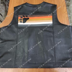 May include: Black leather vest with a bear paw and horizontal stripes in brown, orange, tan, beige, gray, and white. The vest has a classic design with a round neckline and a tailored fit. The back features a decorative panel with the bear paw and stripes.