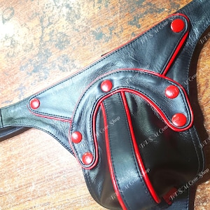 Puede incluir: Arnés de cuero negro con ribetes y detalles en rojo. El arnés presenta costuras rojas y detalles de botones rojos. El producto está diseñado con una forma única y está hecho de un material liso y brillante.