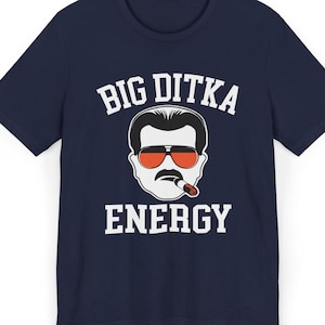 Puede incluir: Camiseta azul marino con un diseño de un hombre con gafas de sol y bigote, con las palabras "BIG DITKA ENERGY" en blanco. El diseño está centrado en la parte delantera de la camiseta.