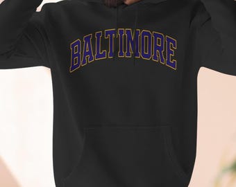 May include: Black hooded sweatshirt featuring the word "BALTIMORE" in purple letters with a gold outline. The sweatshirt has a front pocket and a drawstring hood.