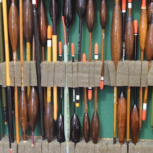 Könnte beinhalten: Eine Sammlung von Angelposen in verschiedenen Formen und Größen, präsentiert in einer Holzkiste mit grünem Filzfutter. Die Posen bestehen aus Holz und Kunststoff mit orangefarbenen, weißen und schwarzen Akzenten. Einige haben eine Tropfenform, andere sind zylindrisch.