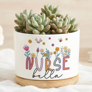 Könnte beinhalten: Ein weißer Keramik-Blumentopf mit einer Sukkulentenpflanze. Der Topf trägt das Wort "NURSE" in bunten Buchstaben, darunter "bella" in Schreibschrift. Blumen- und Insektenillustrationen verzieren den Topf. Der Topf steht auf einem Holzsockel.