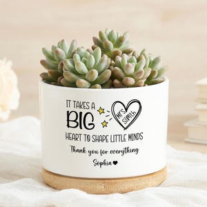 May include: A white ceramic plant pot with a succulent. The pot has the text "IT TAKES A BIG HEART TO SHAPE LITTLE MINDS" and "Thank you for everything Sophia" in black. It sits on a wooden base.