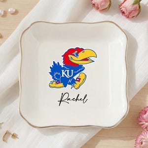May include: A white ceramic trinket dish with a gold rim, featuring a colorful Jayhawk mascot graphic and the name "Rachel" in script. The dish is square with scalloped edges.