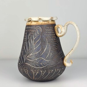 Puede incluir: Una taza de cerámica hecha a mano con un cuerpo azul-gris oscuro y un asa color crema. La taza presenta un diseño de hojas en relieve y un borde decorativo. El asa tiene una forma curva única.