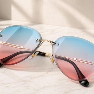 May include: Rimless sunglasses with gradient lenses transitioning from blue to pink. The sunglasses have a gold-colored metal frame and black temples. The lenses are oval-shaped and the sunglasses are resting on a white surface.