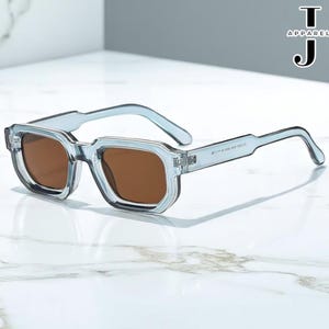 Op de afbeelding: Lichtblauwe vierkante zonnebril met bruine glazen. De bril heeft een modern design met een transparant montuur. De tekst "APARELBYJOSEPH" is rechtsboven zichtbaar. Op een marmeren achtergrond.