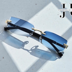 May include: Rimless sunglasses with gradient blue lenses and gold-colored accents. The temples are black. The text "TTT APPARELBYJOSEPH JU" is in the upper right corner. The sunglasses are casting a shadow on a light-colored surface.