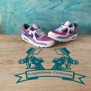 May include: Pair of custom sneakers with white, pink, purple, and teal accents. The shoes feature a star pattern on the side panels and a white sole. The Nike swoosh is purple. The shoes are displayed on a wooden surface.