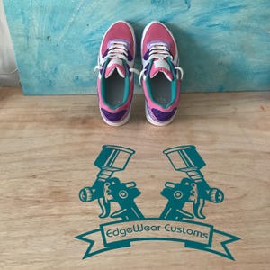 May include: Pair of pink, purple, and white sneakers with white laces. The shoes have a teal interior and accents. The sneakers are displayed on a wooden surface with a teal graphic of two spray guns and the text "Edgewear Customs".