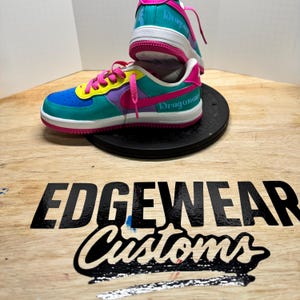 Pu&ograve; includere: Sneakers personalizzate con una vivace combinazione di colori. Le scarpe presentano dettagli blu, verde acqua, viola, rosa e gialli. Il testo "Dragonair" &egrave; visibile sul lato. Le scarpe sono su una piattaforma nera, con il logo "EDGEWEAR Customs" in primo piano.