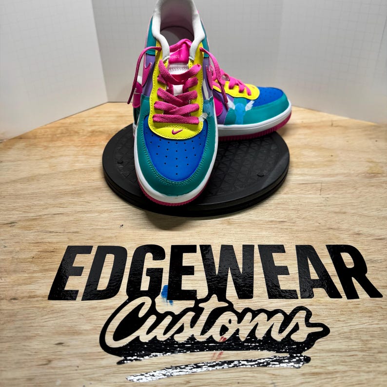 Pu&ograve; includere: Un paio di sneakers personalizzate con una vivace combinazione di colori. Le scarpe presentano una punta blu, lati verde acqua e dettagli gialli. Lacci rosa e uno swoosh bianco completano il design. Le scarpe sono su una piattaforma nera.