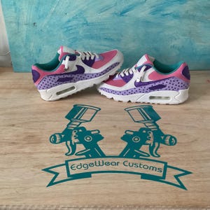 May include: A pair of custom-painted sneakers in pink, purple, white, and teal. The shoes feature a star pattern on the purple sections and a white sole. The logo "EdgeWear Customs" is visible on the wooden surface.