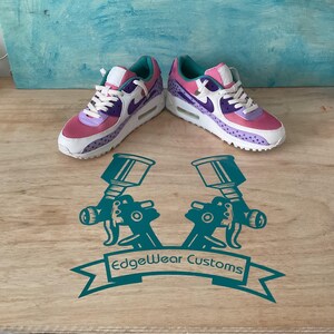 May include: A pair of custom sneakers in pink, purple, and white, with a teal accent. The shoes have a dotted pattern and the Nike swoosh logo. The shoes are displayed on a wooden surface with the text "EdgeWear Customs".
