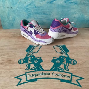 May include: A pair of custom-painted sneakers in pink, purple, and white. The shoes feature a star pattern and the Nike swoosh logo. The soles are white, and the laces are white. The shoes are on a wooden surface with a teal graphic that says "EdgeWear Customs".