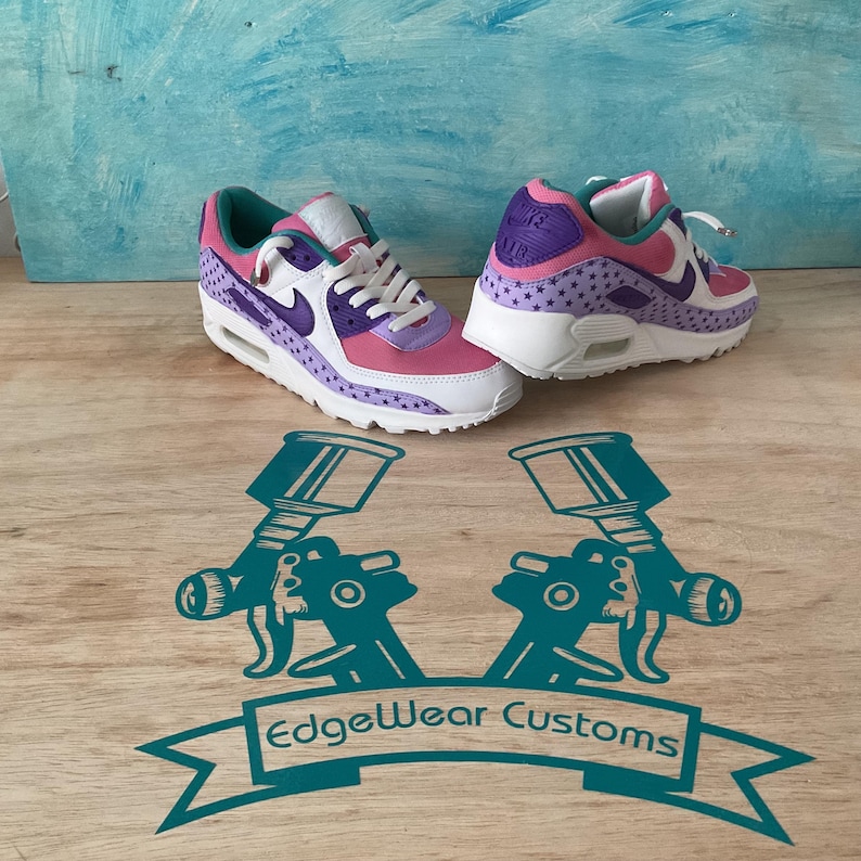 May include: A pair of custom-painted sneakers in pink, purple, and white. The shoes feature a star pattern and the Nike swoosh logo. The soles are white, and the laces are white. The shoes are on a wooden surface with a teal graphic that says "EdgeWear Customs".