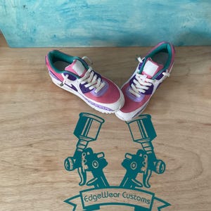 May include: Pair of pink, purple, and white sneakers with white laces. The shoes have a teal interior and accents. The sneakers are on a wooden surface with a teal and blue background. The logo "EdgeWear Customs" is at the bottom.