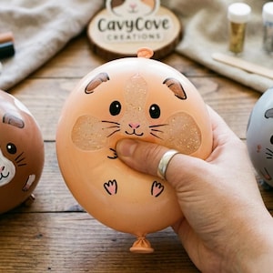 Może przedstawiać: Trzy okrągłe, przypominające balony zabawki antystresowe w kształcie świnek morskich. Jedna jest brązowa z białymi znaczeniami, druga brzoskwiniowa z brokatowymi policzkami, a trzecia szara z ciemnymi plamkami. Każda ma namalowany pyszczek i uszy.