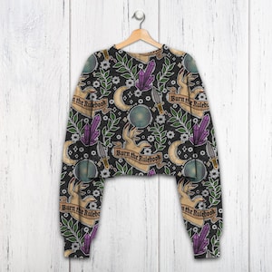 K&ouml;nnte beinhalten: Schwarzes, gecropptes Sweatshirt mit einem sich wiederholenden Muster aus H&auml;nden, Kristallen, Monden, Dolchen und floralen Elementen. Der Text "Burn the Rulebook" befindet sich auf Bannern. &Auml;rmel und Saum sind schwarz.