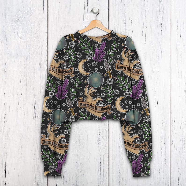 K&ouml;nnte beinhalten: Schwarzes, gecropptes Sweatshirt mit einem sich wiederholenden Muster aus H&auml;nden, Kristallen, Monden, Dolchen und floralen Elementen. Der Text "Burn the Rulebook" befindet sich auf Bannern. &Auml;rmel und Saum sind schwarz.