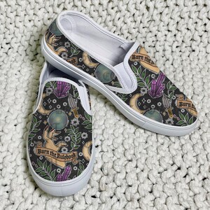 K&ouml;nnte beinhalten: Paar Slip-On-Schuhe mit schwarzem Hintergrund und einem sich wiederholenden Muster aus Monden, Kristallen, Dolchen und H&auml;nden. Die Schuhe haben wei&szlig;e Sohlen und den Text "Burn the Rulebook" auf einem Banner.